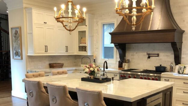 this beautiful kitchen renovation features a large island with a white marble top and a large brown vent hood, luxury appliances and beautiful light fixtures