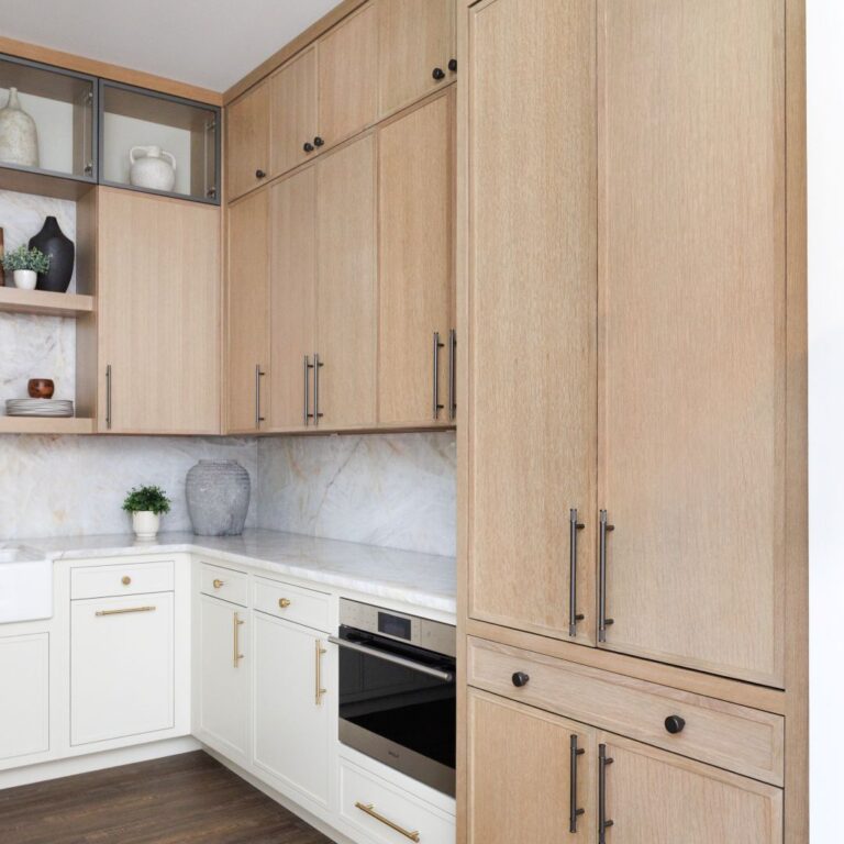 The display features Mouser Eurostyle upper cabinets in Quarter Sawn Oak with a Cadet Stain finish, accented by Top Knobs Burnham hardware in a sleek Ash Gray. Below, Mouser Square Inset lower cabinets in painted Maple with an Eggshell finish pair beautifully with Burnham hardware in a warm Honey Bronze, creating a sophisticated contrast.