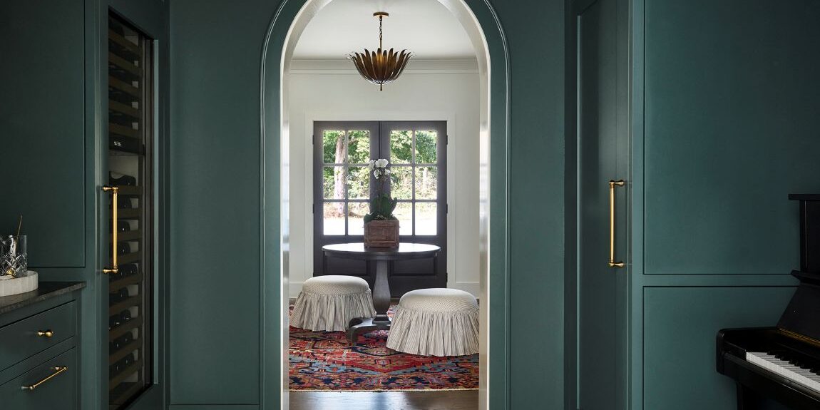 Atlanta home bar and wine storage area featuring dark green custom cabinetry, brass hardware, geometric tile flooring, arched doorway, and moody transitional design details