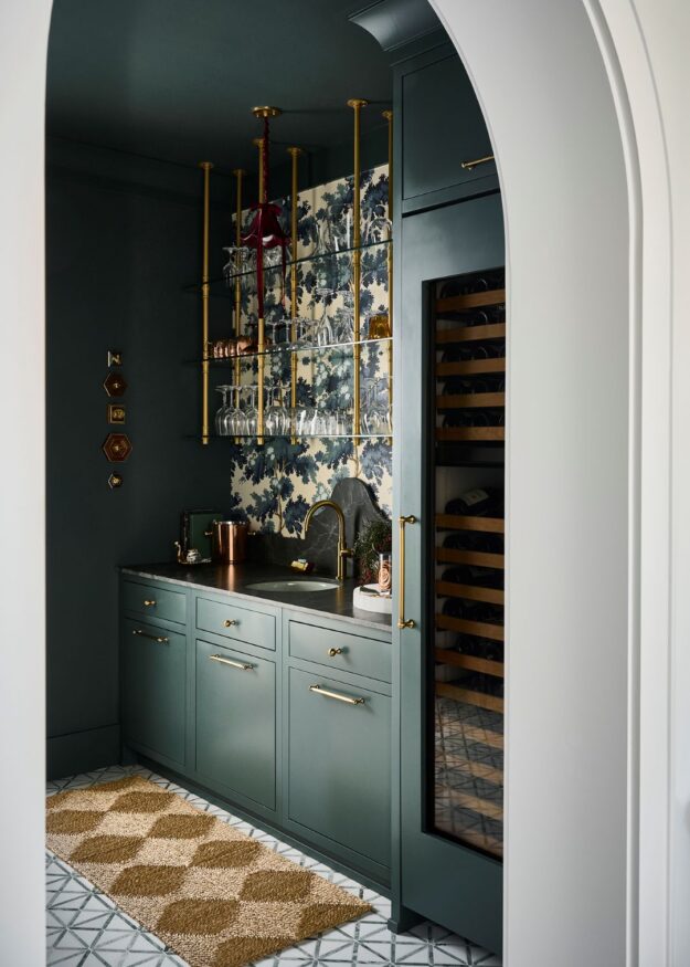 Custom Atlanta bar area featuring dark green cabinetry, brass hardware, floating glass shelves, patterned wallpaper, built-in wine storage, and an arched entryway with dramatic moody design details